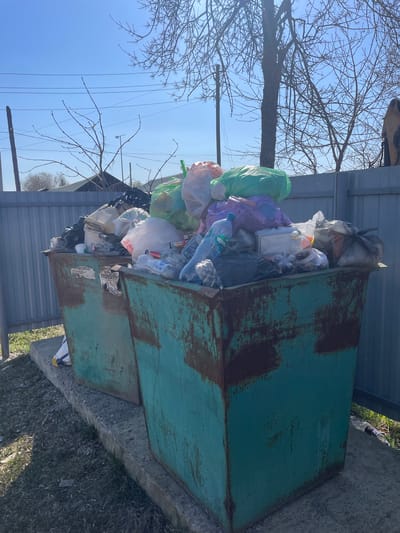 A dumpster is overflowing with garbage bags and other waste. The dumpster is green and rusty, with peeling paint. It is situated on a concrete slab next to a corrugated metal fence. Bare trees are visible in the background against a clear blue sky. Powerlines are also visible in the background.