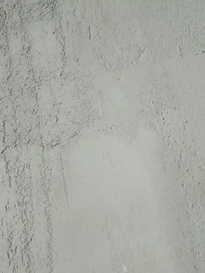 The image is a close-up, full-frame, vertical shot of a grey, textured surface, likely a concrete or stucco wall. The texture is rough and uneven, with visible ridges and small pits scattered across it. There are no people, objects, or discernible actions captured in this abstract textural study. The lighting appears to be diffuse, creating subtle shadows that highlight the unevenness of the surface. The overall impression is one of a simple, unadorned background texture. It is not possible to determine the location in the city of Podmosh'ye, Russia based solely on this image.