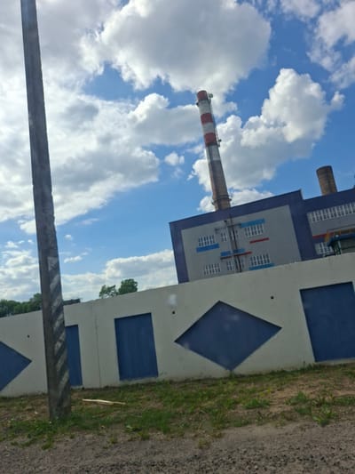 The image shows a building with a tall chimney, likely an industrial facility, against a backdrop of a cloudy blue sky. The building's facade is painted with blue diamond and rectangle patterns. A section of a gray structure is visible in the foreground. The presence of the chimney suggests a power plant or factory. The location of the image is Zhabinka, Belarus.