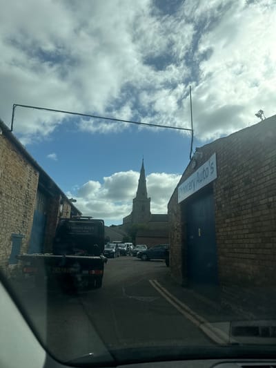 The image depicts a street scene in Thrapston, United Kingdom, with a view framed by brick buildings on either side. A church spire rises in the distance, centered between the buildings and set against a backdrop of cloudy blue sky. Vehicles, primarily vans, are parked along the right side of the street. One van, closer to the camera, has the name "Chancery Auto's" and a phone number displayed on its side. A sign bearing the same name "Chancery Auto's" is visible on the adjacent building. Power lines run across the sky. The image is taken from inside a vehicle, as indicated by the dashboard visible at the bottom.
