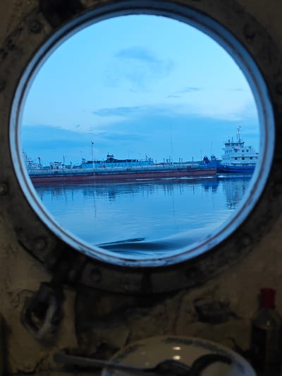 This image shows a view through a ship's porthole, looking out onto a waterway. The sky is a pale blue with scattered clouds, suggesting it is either early morning or late evening. The water is calm and reflects the sky and the ships docked in the distance.
The main subjects are two ships. The closer ship is a long, red-hulled barge. Behind it, a white and blue ship with a tall superstructure is visible, its name clearly marked as "КАПИТАН АЛЕКСЕЕВ" (Captain Alekseev). Several other vessels and structures are also present along the waterfront. The foreground of the image is dominated by the weathered metal frame of the porthole, with some of its fixtures visible, hinting at the interior of a ship. A small bottle with a red cap is visible in the bottom right corner, adding a domestic detail to the scene.
The scene appears to be a harbor or a dock. The muted colors and calm water evoke a sense of tranquility. The presence of docked ships suggests a working port, possibly in a cold climate given the overcast sky. The name on the ship, "Captain Alekseev", and the location context of Vorogovo, Russia, place the scene in a specific geographical and cultural setting. The overall mood is one of quiet observation, with the porthole acting as a frame that isolates and focuses the viewer's attention on the maritime scene.