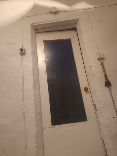 The image depicts an interior scene of a room with white, textured walls. A white door with a dark, rectangular window occupies the center of the frame. The door is slightly ajar.
On the wall to the left of the door, there is a light-colored electrical outlet with a cord running downwards. Above the door, a circular object, possibly a smoke detector or light fixture, is visible. To the right of the door, another electrical outlet is mounted on the wall, and a long, rope-like object with a red and black handle hangs from it. A small, red, round doorknob is visible on the door.
The lighting in the room is dim and appears to come from an unseen source, casting some shadows and highlighting the textured nature of the walls. The overall impression is one of a utilitarian and somewhat aged interior space. There is no indication of people, specific activities, or weather conditions. The location context suggests the scene is in Қаратөбе ауылдық округі, Kazakhstan. No visible text is present in the image.