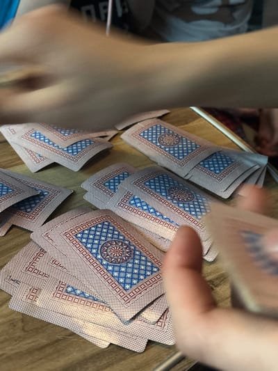 The image depicts an indoor scene in Pervomaiskoe, Russia, focusing on a wooden surface where an activity involving playing cards is underway. Numerous playing cards are scattered across the surface, predominantly in loose piles and individual cards, all displayed with their backs facing upwards. The card backs feature a distinctive blue and red geometric pattern, centered with a star-like motif. Two sets of human hands are visible, both blurred due to motion. In the upper portion of the frame, a blurred forearm and hand are positioned, indicating movement across the scene. In the lower right, fingers from another hand are visible, actively holding or manipulating several cards. The overall impression is one of active engagement in a card game or similar activity.