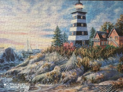 The image depicts a puzzle of a lighthouse and a coastal scene. The lighthouse is tall and striped black and white, with lit windows. To the right of the lighthouse is a red-brick house with a dark roof and lit windows, suggesting it is nighttime or dusk. In front of the lighthouse and house, there are tall grasses and some red foliage. To the left, there is a rough, rocky shore with crashing waves and a sailboat in the distance. The sky is cloudy and shows hints of sunset colors. The image does not provide any information about a specific location in Kugesi, Russia.
