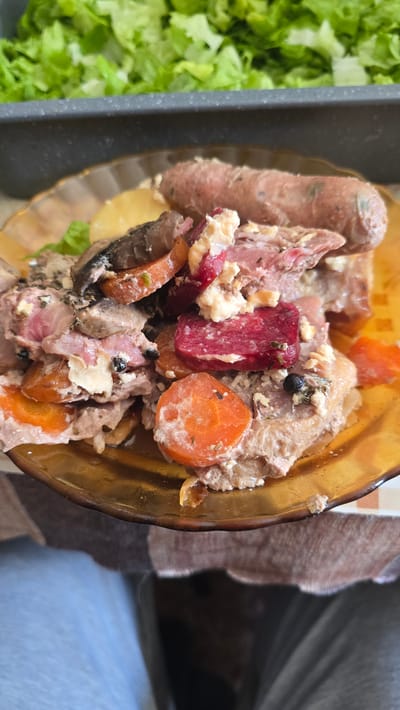 A close-up shot presents a partially consumed dish in a transparent amber bowl, possibly a stew or salad. The ingredients include sausage, meat, carrots, beetroot, pineapple, and lettuce. These items appear mixed and coated in a creamy, possibly cheesy sauce with black peppercorns. A tray of lettuce is visible to the side. The bowl rests on a textured brown and white surface, with a person wearing blue pants or jeans seated nearby. The location in Susam, Bulgaria is unspecified and inferred.
