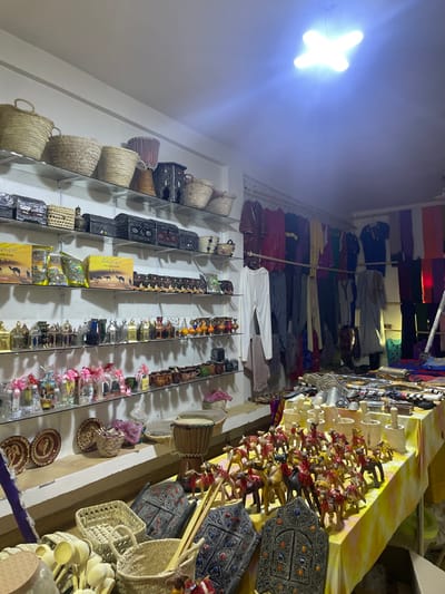 A well-lit shop displays a variety of goods. Woven baskets and embellished boxes line shelves on the left. Clothing hangs above. Further right, shelves showcase small decorative items, including souvenirs. A table displays wooden horse figures and ornamented boxes. The merchandise suggests a market setting or souvenir shop.