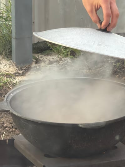 The image shows a close-up, rotated view of a large, dark pot with a light-colored lid being held slightly ajar by a person's hand. Steam is billowing out from the opening of the pot, suggesting that something is being cooked or heated inside. The pot appears to be made of cast iron and has a textured, dark exterior. The lid is textured and appears to be made of a lighter material, possibly aluminum or a similar metal. The hand holding the lid is lightly gripping a knob or handle on its edge.
The scene is set outdoors, with glimpses of dirt, dry grass, and possibly some vegetation around the pot. A gray cylindrical object, possibly part of a structure or support, is visible in the lower left portion of the frame. The lighting suggests it is daytime, but the weather is not explicitly discernible. The overall impression is one of food preparation, likely in an outdoor setting such as a campsite or a backyard. There is no visible text in the image. The setting hints at a rustic or outdoor culinary activity.