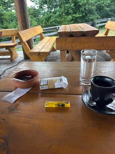 This image captures a close-up view of a wooden table, likely outdoors given the greenery in the background and the rustic wooden furniture. On the table, there are several items: a clear glass filled with water, a black coffee mug with a spoon on its saucer, a pack of cigarettes, a brown ashtray with a cigarette butt in it, a yellow lighter, and a crumpled receipt with a QR code. The wooden furniture, possibly part of a picnic table or seating area, suggests a casual, outdoor setting. The overall lighting and the presence of a coffee mug and cigarettes imply a moment of relaxation, possibly in the morning or afternoon. The text on the cigarette pack is in a foreign language, and the receipt contains pricing information in Euros, with one of the items listed as "Mekića Kula," indicating the location as Mekića Kula in Montenegro. The scene conveys a sense of a brief pause during a meal or a break, with a touch of everyday consumables.