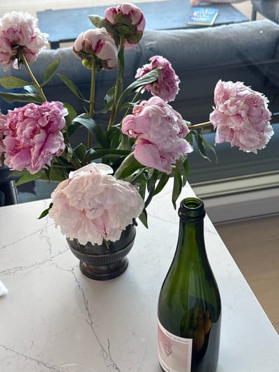 A still life composition features a vase of pink peonies and a green glass bottle with a label on a white marble countertop. The vase, dark and textured, holds several fully bloomed peonies, their petals ruffled and layered in shades of light pink and white, with some deeper pink accents. The bottle is positioned to the right of the vase, its green glass reflecting the surroundings. A light pink label is visible on the bottle, with text that appears to be "Rosé de Mounier" and "CHAMPAGNE." In the background, a blurred blue sofa and a small book with colorful writing are visible, suggesting an indoor domestic setting. The scene is lit with natural light, creating soft shadows and highlights. The image is set in the city of Grad Šibenik, Croatia.