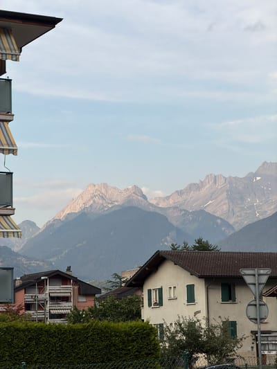 This media file shows a scenic view of mountains behind a residential area. The scene includes a building with a dark tiled roof, light-colored walls, and green shutters on the windows. A traffic sign is visible on the building's exterior. In the foreground, there's a fence and lush green foliage, possibly a hedge. The mountains in the background are rugged and appear to have patches of snow. The sky is mostly clear with some wispy clouds. The lighting suggests it is daytime, possibly late afternoon given the warm tones on the mountains. The overall impression is a peaceful, mountainous landscape in a European setting, likely Monthey, Switzerland.
