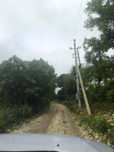 The image depicts a dirt road in a rural setting, likely on the outskirts of Skurdi, Georgia. The road is unpaved and covered with gravel and stones, suggesting it might be a country lane or a service road. It winds uphill, disappearing into the distance. On either side of the road, dense green foliage and trees are visible, with some appearing to be quite tall. A wooden fence runs along the left side of the road for a portion of its length.

In the foreground, the hood of a silver vehicle is visible, indicating the photograph was taken from inside a car. To the right of the road, a utility pole stands, supporting electrical wires that stretch across the frame. The pole is concrete and appears weathered, with some markings or graffiti on it. There is a transformer box attached to the pole. The sky is overcast and appears grayish, suggesting cloudy weather. The overall impression is one of a remote, natural environment.