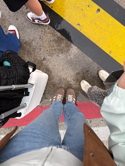 Four individuals stand on a paved surface marked with alternating yellow, black, and red stripes. Each person is visible only from the knees down. One wears blue denim jeans and brown Adidas sneakers. Another is wearing leopard-print pants and white sneakers. The third individual is wearing blue jeans and red and white shoes. The fourth individual is wearing black shorts with white socks and burgundy and white sneakers. A black backpack and a white suitcase are on the ground. The location is Orio al Serio, Italy.