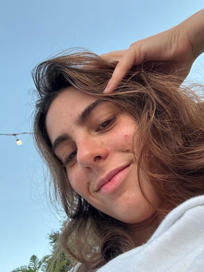 A low-angle, close-up photograph captures a young woman from the chest up, positioned against an outdoor backdrop in Ban Mae Ping, Thailand. The woman has brown, slightly wavy hair, dark eyebrows, and a light complexion exhibiting minor blemishes on her cheeks. Her lips are closed in a slight smile, and she looks directly towards the camera. Her right hand, with manicured fingernails, is visible, with fingers gently inserted into her hair above her forehead on the right side of the frame. She wears a white garment, possibly a hoodie or sweatshirt, visible on her right shoulder and chest. The background features a clear, light blue sky occupying the upper two-thirds of the image. A string of lights with one illuminated bulb is visible in the upper left corner. Green foliage, appearing to be tree branches, is partially visible in the lower left background. The lighting suggests late afternoon or early evening.