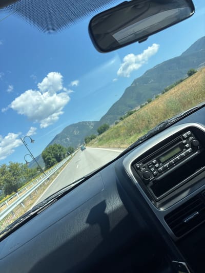 The image is a photograph taken from the interior of a car, looking forward through the windshield. The scene depicts a clear, sunny day with a bright blue sky dotted with fluffy white clouds. In the distance, rolling green hills and mountains form the backdrop, with dry, yellow-brown grass covering the lower slopes, suggesting a rural or semi-rural environment. A paved road stretches into the distance, curving slightly to the left, with a white line marking its edge. A silver guardrail runs alongside the road. Two black lampposts with decorative tops are visible on the left side of the road, near some green trees. A faint blue car can be seen driving ahead on the road.
Inside the car, the dashboard is visible in the foreground. A black car radio with a digital display and various buttons is centrally located on the dashboard. The reflection of the sky and clouds is visible in the car's rearview mirror, which is mounted at the top of the windshield. The windshield itself shows a faint pattern of dots along the top edge, typical of car windshields. The overall impression is of a scenic drive through a mountainous, possibly agricultural landscape. There is no indication of the city of Tulovo, Bulgaria in the image itself.