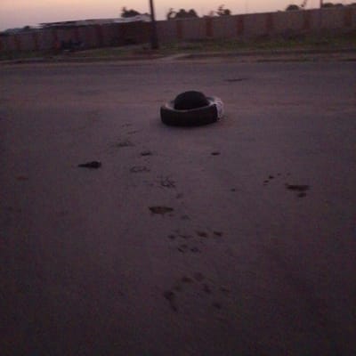 A tire lies on a dark, paved surface, possibly a road, in Wuro Allahamdu, Nigeria. The sky is a dusky, pale orange, suggesting dawn or dusk. In the background, a low, dark structure with a fence can be seen, with some vegetation and trees behind it. The ground in the foreground shows some indistinct marks and patches, possibly footprints or debris, and a small, dark object is visible to the left of the tire. The overall scene is dimly lit and somewhat blurry.