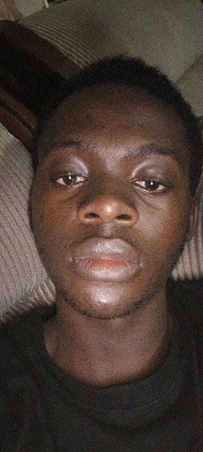 The image displays a close-up, head-and-shoulders portrait of a young adult male with dark skin, positioned in Uweyoba, Nigeria. The individual, centered in the frame, gazes directly forward with a neutral expression. His eyes are open, and his lips are full, slightly parted, showing a reddish tint. Short, dark, curly hair is visible, and faint stubble is present on his chin and upper lip. He wears a dark, possibly black, crew-neck garment. The background consists of a textured, muted brown or grey fabric, possibly upholstery, indicating an indoor setting. The lighting is low, contributing to visible image noise across the scene. No discernible actions or interactions beyond posing for the photograph are evident.