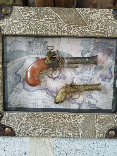 The image shows a framed display containing two toy pistols against a background of a world map. The larger pistol, made of a material that looks like a dark-colored metal with a faux wood handle, is placed above a smaller gold-colored pistol. The frame itself has a faux leather look, with rivets at the corners. The map background includes the text "Via Vee World Classic" in an ornate circle at the top left and "Via Vee world" in another circle near the bottom right. Geographic locations, such as "OCEANUS CHINENSIS" (South China Sea), and other place names, like "Spagna" and "Philippin" are also discernible. The picture appears to be taken on a white or light grey surface, possibly an outdoor wall. There is no indication of a location in Padarsko, Bulgaria within the image.
