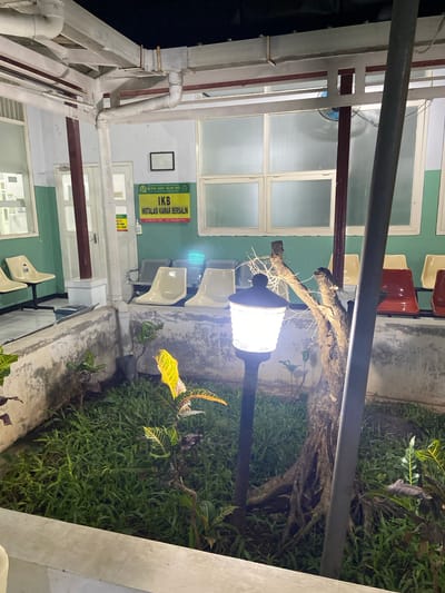 This media file depicts an outdoor waiting area at a facility, possibly a hospital or clinic, in Margoyoso, Indonesia. The scene is lit by a warm-toned outdoor lamp, suggesting it is either dusk or nighttime. Greenery, including grass and potted plants with variegated leaves, surrounds the waiting area.
The waiting area itself features several rows of seating. Some chairs are simple, molded plastic in a light beige color, while others are darker, possibly red or maroon, and appear to be made of metal and plastic. The architecture suggests an open-air or semi-enclosed space with a roof supported by white pillars and metal beams. Visible pipes run along the exterior wall.
A sign in Indonesian is prominently displayed on the green wall. It reads: "RUMAH SAKIT ISLAM PATI" at the top, followed by "IKB INSTALASI KAMAR BERSALIN". Below this, there is smaller text with a phone number and email address. Another smaller yellow sign is partially visible with the word "MAAF".
The overall impression is one of a functional waiting space designed to be illuminated and accessible during darker hours, with a touch of natural landscaping. The presence of the sign for "INSTALASI KAMAR BERSALIN" (Delivery Room Installation) strongly suggests a medical facility, specifically a maternity ward. The lighting creates a somewhat subdued and peaceful atmosphere.