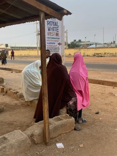 Three women in hijab sit near a bus stop. One wears pink, another brown, and a third white. A sign for "Royal White Guest Inn" advertises "Accommodation, Refreshment, Luxury, Comfort" and a phone number. People are walking in the background. The area appears dusty, with a road in the distance. The bus stop's structure is simple, with a roof supported by wooden beams and concrete blocks.