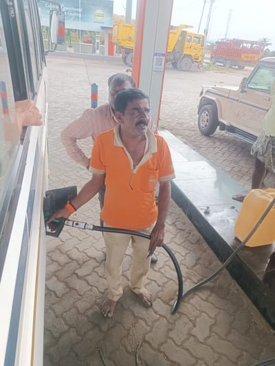At a gas station in Sugalimetta, India, a man in an orange polo shirt and tan pants is filling a vehicle with fuel. He is holding a green fuel nozzle connected to a hose, which is inserted into the vehicle's fuel tank. Another man, wearing a light brown shirt, stands behind him, partially obscured. In the background, a yellow dump truck and a red truck are visible, along with a beige SUV. The ground is paved with interlocking concrete blocks. A large yellow jerrycan is placed on a low stone structure to the right of the frame. The sky appears overcast.