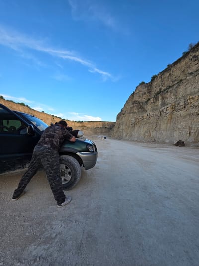 A person wearing camouflage clothing is leaning over the hood of a dark-colored SUV in a quarry. The person is looking down at something on the hood. The SUV is parked on a dirt road in a quarry with tall, layered rock walls on the right side of the image and a blue sky with some clouds above. The quarry appears to be an industrial or mining site, possibly located in Suvorovo, Bulgaria, given the context. There is a large piece of machinery or equipment, resembling a bucket or scoop, visible in the distance on the right side of the road.