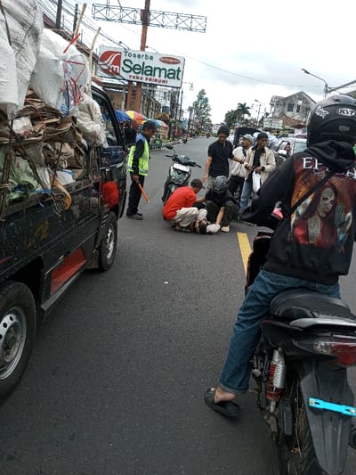 The image depicts an urban street scene in the city of Taohom, Laos, under an overcast sky. In the mid-ground, a small gathering of approximately seven individuals surrounds a person lying prone on the asphalt road. Two individuals, one in a red t-shirt and another in a dark t-shirt and helmet, are kneeling beside the prone person, appearing to provide assistance. Adjacent to this group, a man in a blue shirt and yellow-green reflective vest stands, holding an orange baton, positioned near a heavily loaded black pickup truck. The truck's bed is overflowing with white sacks and cardboard, secured by ropes. Further back, a light-colored scooter is parked. In the immediate foreground, facing away from the scene, an individual is seated on a black motorcycle, wearing a black helmet, blue jeans, black flip-flops, and a black hoodie with an "EPICA" graphic featuring a woman's face. Along the street in the background, a row of multi-story buildings is visible, topped by a prominent sign displaying "Toserba Selamat" and "TOKO PRIBUMI" in red and green text. The road surface is dark asphalt with a visible yellow line. Utility poles and overhead wires are present throughout the background.