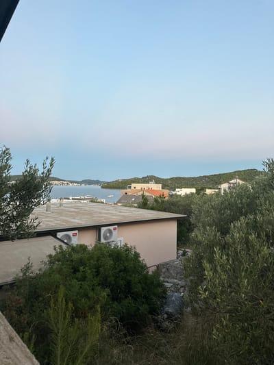 The image captures a panoramic, elevated view of a coastal town identified as Betina, Croatia, during either dusk or dawn. The sky, occupying the upper two-thirds of the frame, transitions from a light blue overhead to a subtle pinkish-blue gradient near the horizon.
Below the horizon, a calm body of water, indicative of a bay, stretches across the mid-ground, dotted with several small white boats. This bay is bordered by low, undulating hills covered in dense green Mediterranean vegetation.
In the immediate mid-ground, a single-story building with light pinkish-beige walls and a flat, light grey roof is prominent. Two white outdoor air conditioning units, bearing the "TCL" logo, are mounted on its visible side wall. Beyond this structure, various residential buildings with light-colored facades and tiled roofs (some orange-red, some grey) are nestled among the green hillsides.
The foreground features natural vegetation, including portions of an olive tree with grey-green leaves on the left, and a dense cluster of olive trees and other shrubs on the right. Below the prominent building, the terrain consists of dark green shrubs, lighter, dried grasses, and exposed rocks. A dark, partially obscured cylindrical object lies among these natural elements in the lower-right mid-ground. No human figures or discernable activity are depicted.