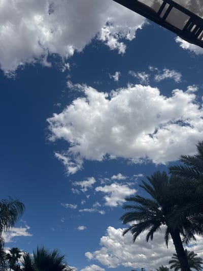 The image shows a view looking upwards towards a bright blue sky filled with numerous white clouds of varying sizes and shapes. Palm trees are visible in the top right and bottom right corners of the frame. A dark structure, possibly a building element or a section of scaffolding, is present in the upper left corner. There are no people visible in the image. The location is within the city of Coachella, United States of America.
