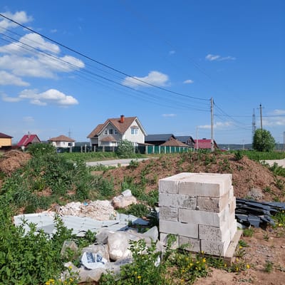 The image shows a construction site in Bol'shaya Mos', Russia. A stack of concrete blocks is visible in the foreground, along with debris like scraps of metal and plastic, and overgrown vegetation. Beyond the construction site, houses with red and brown roofs, and a light-colored house with a brown roof can be seen. Overhead power lines stretch across a blue sky with scattered clouds.