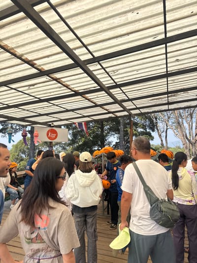 A group of people stands outdoors, some wearing orange helmets. One man wears a white shirt and gray pants, a gray bag slung across his shoulder. A woman stands in front, wearing a gray t-shirt and glasses. A Thai flag is visible in the background, next to a sign with a red logo. An open-roofed metal structure with a corrugated ceiling covers part of the area where the group is standing. Trees and the sky are visible in the background.