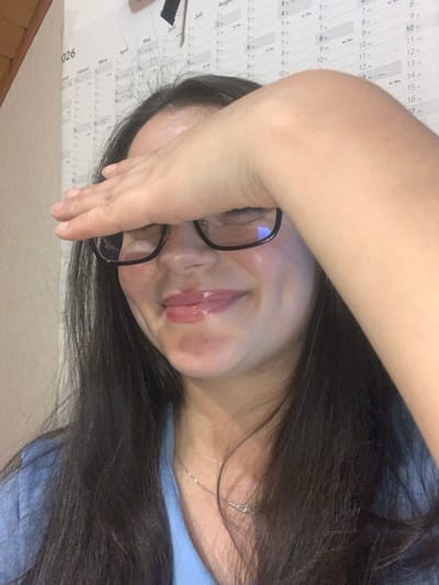 A woman wearing glasses rests her forehead against her hand, partially obscuring her face. She has long, dark hair and glossy lips. A silver necklace is visible at her throat. In the background, a calendar is affixed to the wall, with visible dates and months printed on it, though the calendar is out of focus. The overall composition is a close-up, casual snapshot.