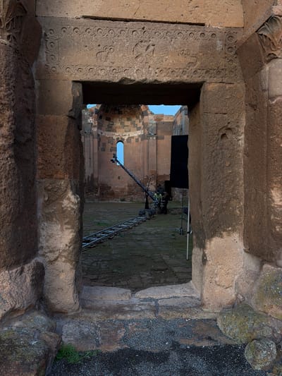 The image shows a view through a stone window or opening in what appears to be an ancient structure in Anipemza, Armenia. The opening is framed by intricately carved stone on the left side, while the stones on the right exhibit a rougher texture. Through the opening, one can see another part of the structure, which appears to be a church or cathedral, with a dome or arched roof visible. A telescopic crane or platform is positioned outside the opening, seemingly for maintenance, filming, or research purposes. Two people are visible operating the crane. The ground outside the opening appears to be a mix of gravel and vegetation.
