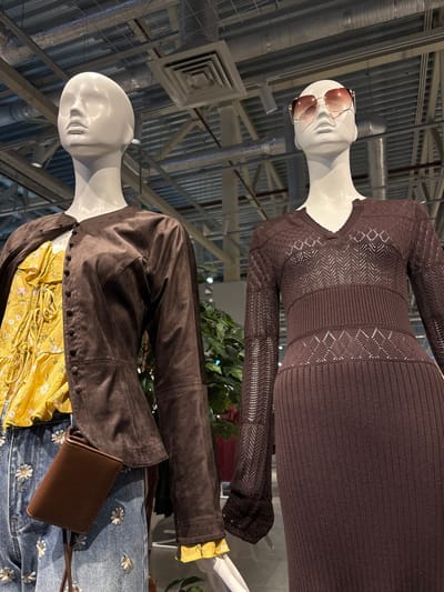 Two mannequins are on display. The top mannequin wears a brown, long-sleeved knitted dress and sunglasses. Below, another mannequin is dressed in a yellow blouse under a brown suede jacket. This outfit is paired with jeans decorated with small daisies and a brown, crossbody bag. The background shows a modern, industrial space with metal beams and ventilation systems.