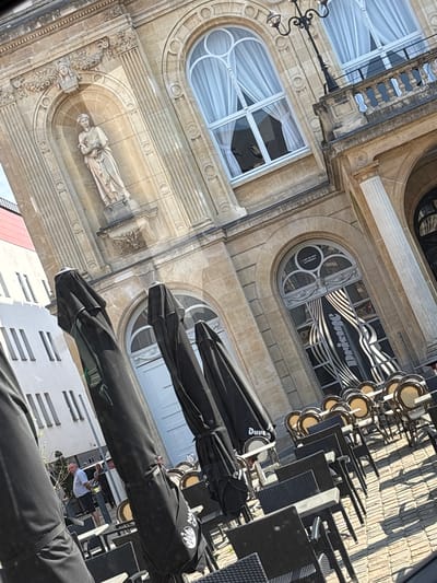 The image shows an outdoor cafe scene in Namur, Belgium. The cafe is situated in front of an ornate, historic building with arched windows and decorative stonework. Several black patio umbrellas are set up, some closed and some partially open. Rows of outdoor tables and chairs are arranged on a cobblestone patio. In the background, people are visible, including a man in a light-colored shirt and dark shorts standing near some planters, and another person in the distance. The architecture suggests a location in the city center, possibly near a prominent square or street.