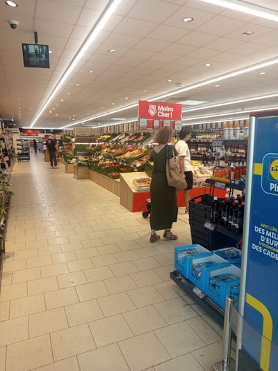 The image shows the interior of a grocery store, likely a Lidl supermarket, located in Laxou, France. The floor is tiled with large, light-colored square tiles. Overhead, fluorescent lighting illuminates the space. In the foreground on the right, a blue promotional sign is partially visible, featuring yellow and white text. Below this sign, blue cardboard boxes containing packaged goods are displayed on a shelf. A black plastic crate containing several dark bottles is positioned above these boxes.
In the midground, a woman wearing a long, dark green dress and carrying a woven bag is walking away from the camera. To her left, a man in a white t-shirt and black shorts stands near a display of produce. Behind them, more shelves are stocked with various food items, including fruits, vegetables, and bottled products. Further in the background, other shoppers are visible, and a section of the store with a red sign reading "Offres Spéciales" (Special Offers) is present. On the ceiling, a security camera and a small display screen are mounted.