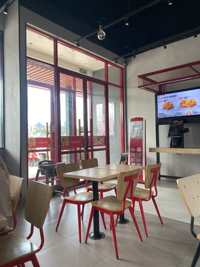 This media file is a photograph of the interior of a fast-food restaurant, possibly a KFC, in Lhoksukon, Indonesia. The image is taken from a high angle, looking down at a seating area with several wooden tables and chairs. The chairs have red metal legs and backs, and the tables appear to be made of light-colored wood with dark metal bases.
On the left side of the image, a large window with red framing looks out onto a street or outdoor area where a red motorcycle is visible. The window has horizontal blinds or louvers on the inside. Signs with text in Indonesian, such as "JAJAH" and "It's Quality Time," are visible through the glass. An "EXIT" sign with green lettering is also present.
In the background, a digital menu board displays images of fried chicken and prices in Indonesian Rupiah. Text on the menu board reads "A LA CARTE CHICKEN" and "MAKIN LENGKAP." A red vending machine labeled "New Release" is also visible. The overall lighting suggests it is daytime, and the atmosphere appears to be that of a casual dining establishment. The image is oriented vertically.