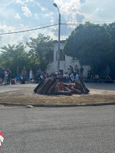 The image, oriented incorrectly, depicts an outdoor community gathering around a large bonfire under a bright, cloudy sky. When correctly oriented, the scene shows a wide asphalt road or paved area in the foreground on the right, separating a grassy verge on the far right from the main event area.

In the mid-ground, a substantial bonfire made of stacked logs is actively burning, with visible flames and glowing embers. Numerous people are gathered around the bonfire, observing the flames. Many are seated on portable folding chairs, while others stand. The crowd consists of individuals of various ages, dressed in casual attire. They appear engaged and observant of the fire.

In the background, behind the gathering, an old, light-colored building with dark window openings is visible, suggesting a traditional or weathered structure. Green trees with dense foliage are present throughout the background and on the left side of the frame, providing shade. A utility pole with wires and a streetlamp is also visible on the left. The sky is bright blue with scattered white cumulus clouds, indicating a clear, sunny day, likely late afternoon or early evening, given the bonfire.

This event, centered around a large bonfire in Balgari, Bulgaria, strongly suggests the preparation or early stages of the traditional Nestinarstvo (fire-walking) ritual, a prominent cultural practice in the village. A small red and white striped object, possibly a flag, is partially visible in the bottom right corner (lower left when rotated correctly). No discernible text is visible in the image.