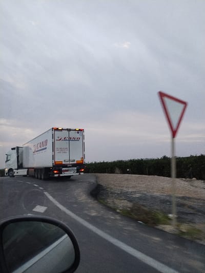 A white semi-trailer truck with "J. CANO" branding is on a gray road. A yield sign is prominently visible on the right side of the road. The roadside is lined with greenery and low walls. A side mirror of a car captures part of the frame, suggesting a driver's perspective. Overcast sky fills the background.