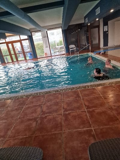 The image depicts an indoor swimming pool facility located in Odorheiu Secuiesc, Romania. A rectangular pool with blue water, exhibiting surface ripples, occupies the central and left portions of the frame. The pool is bordered by reddish-brown square floor tiles and a mosaic-patterned drainage edge. Three individuals are partially submerged in the pool on the right side. In the foreground, an individual with dark hair, appearing female, faces right. Behind her, an individual with a light/bald head, appearing male, faces slightly left. Further to the left and rear, a third individual with light hair, appearing female, faces right. A metal handrail extends into the pool near the three individuals. In the far left background, a partially visible individual, appearing male, stands outside the pool area beyond large glass doors and windows, which offer a view of green hedges and two white chairs. The interior features a light-colored ceiling supported by blue structural beams, equipped with vents and recessed lighting. On the tiled deck to the far right, blue mats and various personal items are visible. The extreme foreground contains the edge of a dark, wicker-patterned piece of furniture.