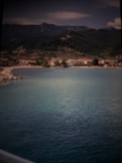 The image depicts a coastal scene with a body of water in the foreground and a town nestled against a mountainous backdrop in the distance. The water appears calm and reflects the muted colors of the sky and land. The town is characterized by buildings with reddish-brown roofs, suggesting a Mediterranean architectural style. A curved, sandy beach is visible along the coastline, bordered by a pier or breakwater. The mountains behind the town are covered in dense vegetation, and their peaks are shrouded in soft, diffused light. The overall impression is one of a serene and picturesque landscape. The provided crops offer closer views of the mountainous terrain and the town's coastal area, reinforcing the impression of a natural and inhabited coastal environment. The image's blurriness suggests it may have been captured with a slow shutter speed or from a moving platform.