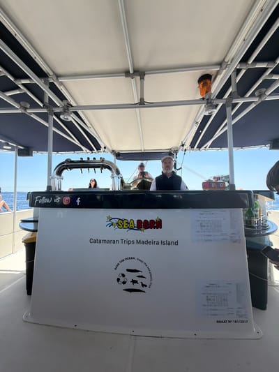 The image depicts an outdoor scene on a boat, featuring a white structure with the text "SEA BORN" and "Catamaran Trips Madeira Island." The structure also has social media icons, suggesting it is promoting a tourism-related business. People are visible in the background, indicating they are passengers or crew on the boat. The image captures a portion of the ocean and clear sky above, suggesting a coastal setting. Additionally, there is a "TIP BOX" on the structure and a sign that says “SAVE THE OCEAN. Keep the sea plastic free”. Based on the information, the location is likely in Madeira Island, Portugal. The city name, Caniço, Portugal, is not evident within the image.