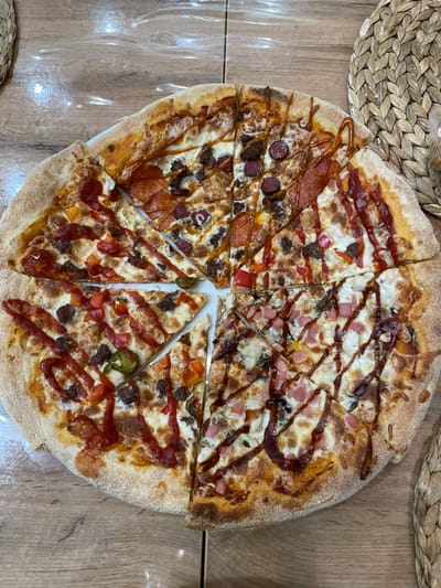 The image shows a sliced pizza with multiple toppings on a wooden surface. The pizza appears to have been cut into several uneven slices. Two place mats are partially visible around the pizza. The location is Лесной Пейзаж Кп (шарапово), Russia. No people are visible.