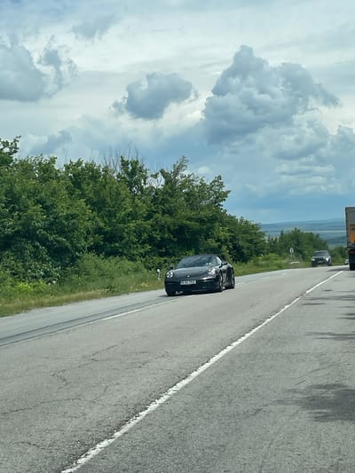 The image depicts a black Porsche 911 driving on a two-lane road, likely in a rural area. The car's license plate reads "B 93 TRZ." The road is flanked by green trees and vegetation. In the distance, a landscape with a horizon line is visible. The sky is filled with white and gray clouds. A second vehicle can be seen further up the road. The image suggests it was taken in daylight under a partly cloudy sky. The location is possibly near Osenets, Bulgaria, given that the license plate is likely a Bulgarian plate.