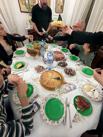A group of people are gathered around a table laden with food and drinks in a brightly lit indoor setting. The table is covered with a white tablecloth adorned with floral-patterned plates and napkins. A variety of food items, including cheese, salami, a large pie, and cake, are spread across the table. There are also bottles of water and a bottle of sparkling wine or champagne. Several people are actively engaging in serving or pouring drinks, while others are seated at the table.

A woman with dark hair is raising a glass in one hand while a man with a beard is pouring sparkling wine into it with the other. To the right, a person with a striped shirt has a phone resting beside their plate.

The location appears to be a dining room, suggested by the presence of furniture, artwork on the walls, and curtains. There is no explicit evidence to confirm the location within the city of Tarquinia, Italy.