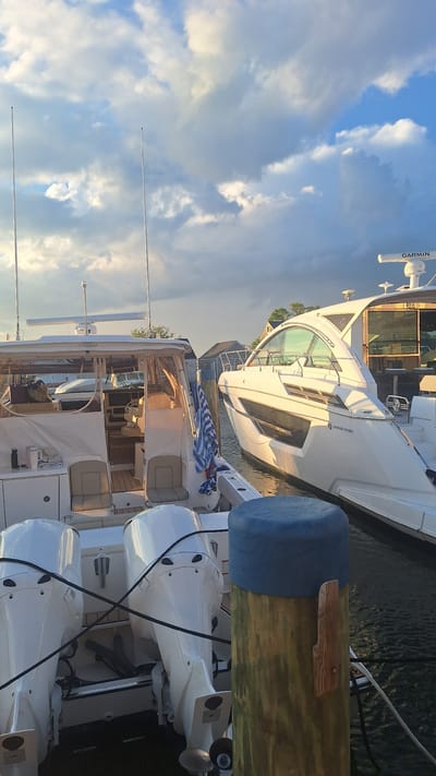 The image captures a close-up view of two boats docked in a harbor, likely in Nantucket, Massachusetts, USA, during the late afternoon or early evening. The sky above is filled with dramatic, cumulus clouds, some illuminated by the warm, setting sun, casting a golden glow on the scene.
In the foreground, the stern of a white boat is visible, equipped with twin white outboard motors. The boat appears to be moored to a wooden pilothad a blue protective cover. A black rope runs diagonally across the outboard motors.
To the right of this boat is a sleek, white motor yacht with a distinctive, curved windshield and dark tinted windows. The yacht is also docked and appears to be well-maintained. A radar dome labeled "GARMIN" is visible on the roof of a structure attached to the yacht.
Between the two boats, a blue and white striped towel is draped over a wooden post. In the background, several houses with light-colored siding and dark roofs are visible, suggesting a waterfront residential area or a marina. The water is dark and calm, reflecting the sky and the boats.
There are no people visible in the image. The overall impression is one of a peaceful, serene evening at a harbor.