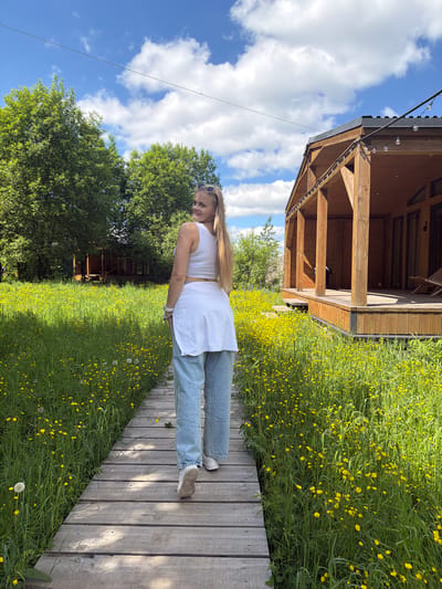 The image, taken in Vozhoiskii, Russia, captures a bright outdoor scene on a sunny day. A young woman with long blonde hair, wearing a white sleeveless crop top, light blue jeans, and light-colored sneakers, is the main subject. She stands on a wooden walkway, looking slightly to her left with a relaxed expression and a subtle smile, her sunglasses resting on her head.

The walkway stretches from the foreground into the distance, running alongside a vibrant field of lush green grass densely covered with small yellow flowers, resembling buttercups. Sunlight illuminates the scene, casting subtle shadows on the wooden planks of the path and the surrounding foliage.

In the background, a rustic wooden gazebo or open-sided structure with a dark roof stands amidst the greenery. Decorative string lights are visible along the eaves of the gazebo. Beyond this structure and the field of flowers, a line of dense green trees frames the horizon, beneath a clear blue sky dotted with fluffy white clouds. The overall impression is one of a peaceful and pleasant outdoor setting, likely a park or recreational area, during the daytime.