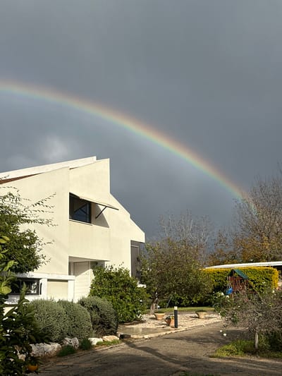 The image depicts a residential scene in Beit Yitshak Sha'ar Hefer, Israel, under a dark, overcast sky with a prominent rainbow spanning across the upper half of the frame. In the left foreground and midground, a two-story, cream-colored house with a modern, angular design is visible. The house features multiple windows, including one on the second floor with an open light-colored awning, and another on the ground floor with dark shutters. The property includes a garden with various green foliage, including bushes, small trees, and potted plants arranged along a paved pathway that curves through the yard. In the midground and background to the right of the house, more trees, some with sparse leaves, are present, alongside a children's playset featuring a slide and a small blue swing, partially obscured by a dense green hedge. No individuals are present or engaged in any actions within the depicted scene.