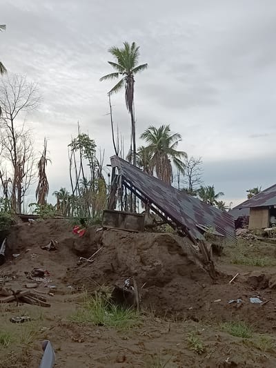 The image shows a scene of destruction in Meunasah Raya, Indonesia. A collapsed corrugated metal roof lies at an angle, partially obscuring a pile of dark, moist earth. Debris, including torn fabric and scattered objects, is visible on the disturbed ground. Tall, slender trees, some bare and others with sparse foliage, surround the area, along with several palm trees. In the background, a partially intact building with a corrugated metal roof stands. The sky is overcast with a dull, cloudy appearance. The overall impression is one of a natural disaster having recently occurred, causing significant damage to structures and the surrounding environment.