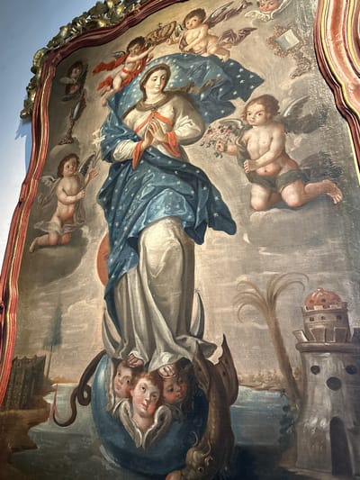This media file captures a detailed religious painting, likely a ceiling fresco or large canvas, within an ornate, gilded frame. The central subject is a female figure, presumably the Virgin Mary, depicted in a celestial setting. She wears a white gown and a flowing blue mantle adorned with white stars, and has a serene expression. Her hands are clasped or holding a small object near her chest. She appears to be standing on a crescent moon or a stylized globe.
Numerous winged putti (cherubs or small angels) surround her. One cherub, positioned above her head, holds a crown. Others are depicted in various poses of adoration or activity, some holding floral bouquets, and one near the bottom left holds a mirror. Below the central figure, a cluster of heads emerges from a cloud-like mass, some looking upwards. Beneath this, a dark serpent is visible, being subdued or crushed, a common symbol associated with the Immaculate Conception.
The painting features a rich, yet aged, color palette dominated by blues, whites, and earthy tones, indicative of Baroque or Colonial art styles. The brushwork suggests a detailed and dramatic composition. In the upper right corner, a structure resembling a fortified building or castle with a dome-like top can be faintly seen in the background. The overall activity portrays the glorification of the Virgin Mary, surrounded by heavenly beings. The painting shows signs of age, including some cracking and darkening of the surface. No discernible text is visible within the artwork itself. The ornate frame suggests this piece is part of a significant architectural or devotional setting, consistent with its location in Tepotzotlán, Mexico.