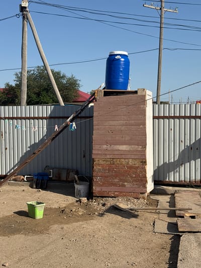 The image depicts an outdoor scene in a rural or semi-rural setting, likely in Аққыстау ауылдық округі, Kazakhstan. In the foreground, a dirt and gravel ground is visible, with a small green plastic bucket in the lower left quadrant. To the right, a wooden shovel lies on the ground, resting on some wooden planks.

Dominating the center of the image is a tall, rectangular structure made of stacked wooden planks, with a white top. On top of this structure sits a large, blue plastic barrel with a white lid. Behind this structure, a corrugated metal fence runs horizontally across the midground, with a section of a wooden utility pole visible to the left, supporting overhead wires. Another utility pole with wires is visible in the background.

To the left of the central structure, a rusty metal ladder leans against the fence. A clothesline is strung across the ladder and fence, with several colorful clothespins attached. A white bucket with some material inside is located to the right of the ladder, near the fence. Various objects, including a blue plastic container and what appear to be metal pipes or tanks, are situated on the ground behind the ladder and fence. The background shows trees and the roofs of buildings, suggesting a residential area. The sky is clear and blue.